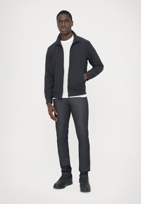 Giacca con zip grigio scuro, t-shirt bianca sotto, jeans di jeans blu, sneakers nere. Tessuto liscio e design aderente.