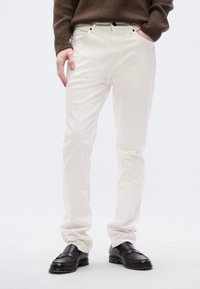 Λευκό βαμβακερό παντελόνι με ίσια γραμμή, που διαθέτει μπροστινή τσέπη, θηλιές για τη ζώνη και ελάχιστη ραφή, σε συνδυασμό με μαύρα δερμάτινα παπούτσια.