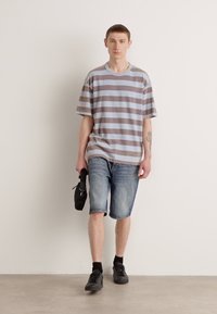 Grå og blå stribet oversized t-shirt, denimshorts, sorte sneakers; model der holder en lille sort taske, mod en neutral baggrund.