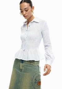 Triibuline hele sinine vormitud särk pepitude äärega ja nööpidega ees, sobitatud teksaseelikuga, millel on suur tasku ja sildike.