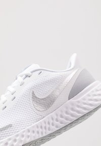 Chaussure de course Nike blanche avec une tige en mesh, une surface texturée, un Swoosh argenté réfléchissant et une semelle intermédiaire ondulée et amortie.