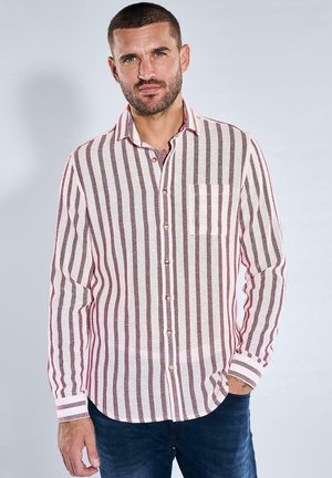 Mann mit kurzen Haaren und Bart, der ein weißes und rot vertikal gestreiftes Hemd und dunkelblaue Jeans trägt, steht mit einer Hand in der Tasche.