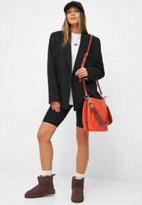 Zwarte dubbelknop blazer over een grijze T-shirt, gecombineerd met zwarte fietsbroek en bruine enkellaarsjes. Geaccentueerd met een oranje handtas en een zwarte pet.