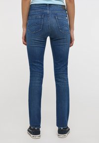 Sötétkék farmernadrág, slim fit fazonban. Hátsó zsebekkel rendelkezik, ívelt varrással és bőr folttal a derékszíjon. Sötét tornacipővel viselve.