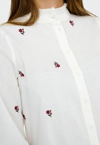 Blusa bianca con bottoni, caratterizzata da piccole ricami floreali rossi e un neckline arricciato, indossata da una persona con capelli castani.