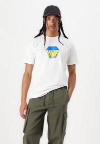 Weißes Baumwoll-T-Shirt mit blauem und gelbem HVF-Logo. Kombiniert mit olivgrünen Cargo-Hosen mit Seitentaschen. Schwarze Cap auf dem Kopf.