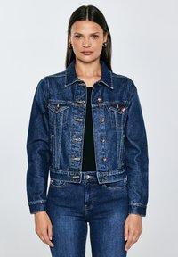 Moteris, vilkinti aptemptą tamsiai mėlyną džinsinę striukę ir derančius džinsus, stovinti atpalaiduotomis rankomis priešais paprastą šviesią foną.