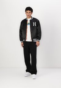 Jachetă bomber neagră din satin cu dungi albe, cu litera "H" pe piept, purtată peste un tricou alb cu un grafic colorat, asortată cu pantaloni negri.