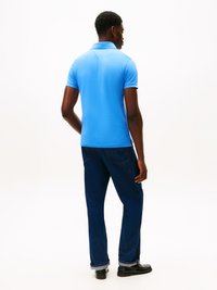 Polo de manga curta em tecido azul brilhante, com colarinho e corte ajustado, combinado com jeans de denim azul escuro com as bainhas dobradas.