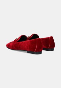 Zapatillas de terciopelo rojo tipo slip-on con un perfil bajo, que presentan una textura suave, punteras redondeadas y detalles decorativos en metal. Suela negra.
