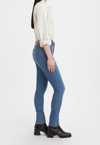 Mujer vestida con una camisa de rayas claras, jeans ajustados azules y botines negros de tacón, de pie contra un fondo blanco liso.