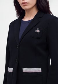 Femme portant un blazer noir avec deux poches inférieures ornées d'un liseré rayé blanc et rose et une petite épingle fleur sur la poche supérieure.