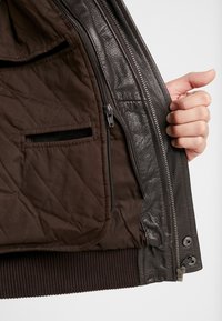 Mano sosteniendo una chaqueta de cuero marrón oscuro abierta que muestra el forro interior acolchado con bolsillos con cremallera y deslizantes, y una cintura acanalada.