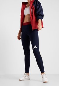 Červená a námořnická větrovka, bílá sportovní podprsenka a námořnické legíny s bílým logem Adidas. Tenisky jsou světle béžové s šedými akcenty.