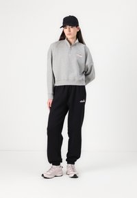 Pelēks zip-up svīteris ar īsu dizainu, melni sporta bikses un melna cepure. Tenisas apavi ir gaiši rozā ar tumšiem akcentiem.