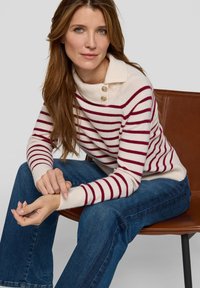 Cremefarbener Pullover mit kastanienbraunen Streifen, gerippter Textur und einem umgeschlagenen Kragen mit zwei goldenen Knöpfen. Kombiniert mit blauen Schlagjeans.