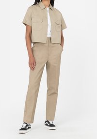 Krátká béžová košile se dvěma předními kapsami, spárovaná s odpovídajícími béžovými kalhotami. Černobílé tenisky dotvářejí tento outfit.