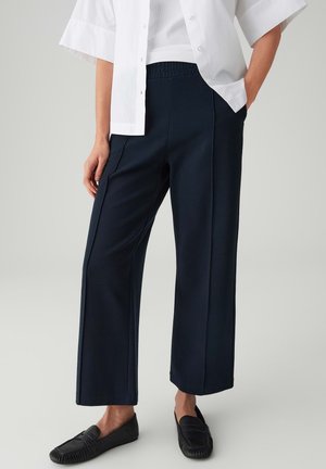 Person trägt schwarze, weit geschnittene Hosen mit vorderen Nähten, weißes Kurzarmhemd, weißes Unterhemd und schwarze Loafer und steht mit einer Hand in der Tasche.