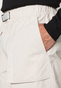 Světlé béžové kraťasy s elastickým pasem a boční kapsou. Tkanina má strukturovaný povrch a na kapse je umístěno bílé logo.