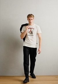 Camiseta blanca con texto gráfico rojo, pantalones negros y zapatos negros. El modelo sostiene una chaqueta negra sobre su hombro, de pie sobre un suelo de madera.