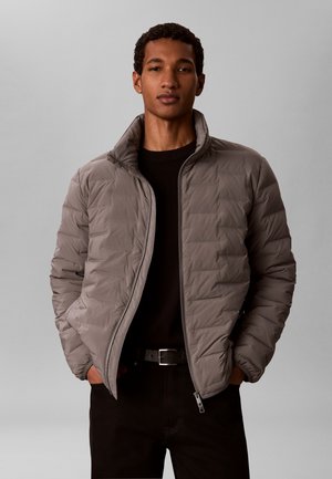 Junger Mann trägt eine graue Steppjacke mit Reißverschluss über einem schwarzen Pullover, die Hände in den Taschen, vor einem einfarbigen hellgrauen Hintergrund.
