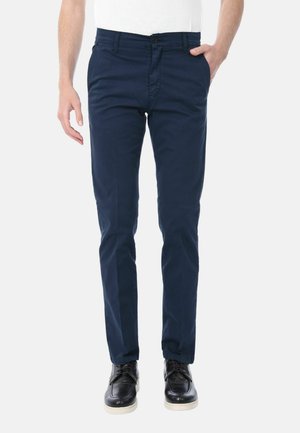 Uomo con pantaloni slim fit blu navy, camicia bianca e scarpe di pelle nere, in piedi con una mano in tasca su sfondo bianco.