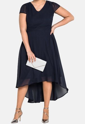 Marineblauwe jurk met korte, doorschijnende mouwen, V-hals en een zoom die bij de voorkant korter is dan bij de achterkant. Gladde stof met een getailleerde taille. Gecombineerd met een zilveren clutch en hakken.