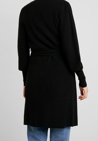 Svart ribbad cardigan med långa ärmar och knytmidja. Texturerat stickat tyg, knälång, stilrent över blå jeans. Enkelt och funktionellt design.