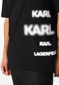 Μαύρη βαμβακερή μπλούζα με λευκό κείμενο σε κουκίδες, "KARL" και "LAGERFELD," τοποθετημένα μπροστά σε διάφορα μεγέθη και στυλ.