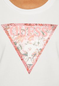 Bela bombažna majica s širokim roza trikotnim grafičnim vzorcem s cvetličnim motivom, logotipom "GUESS" in dekorativnimi žebljički. V središču vprašaj.