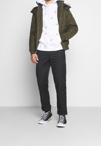 Parka verde con cappuccio, felpa bianca con piccoli loghi stampati, pantaloni scuri e sneaker alte nere. Abbigliamento casual a strati.