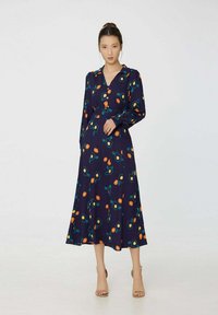 Robe florale marine présentant des manches longues, un col en V et une silhouette fluide. Décorée de fleurs orange et bleu-vert sur un fond sombre.