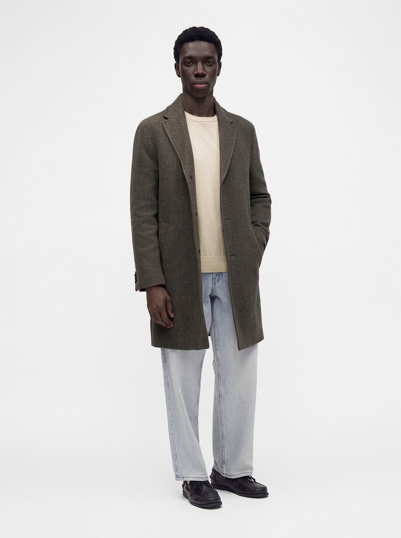 Homme portant un long manteau marron par-dessus un pull beige et un jean bleu clair, debout avec une main dans la poche du manteau sur un fond uni.