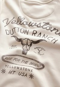 Sweat-shirt beige avec texte imprimé noir, graphique de crâne de taureau et motif décoratif de corde. Le texte comprend "Yellowstone Dutton Ranch" et "MT USA."