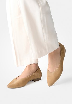 Feet wearing beige suede pointed-toe flat shoes paired with white wide-leg pants against a plain background.