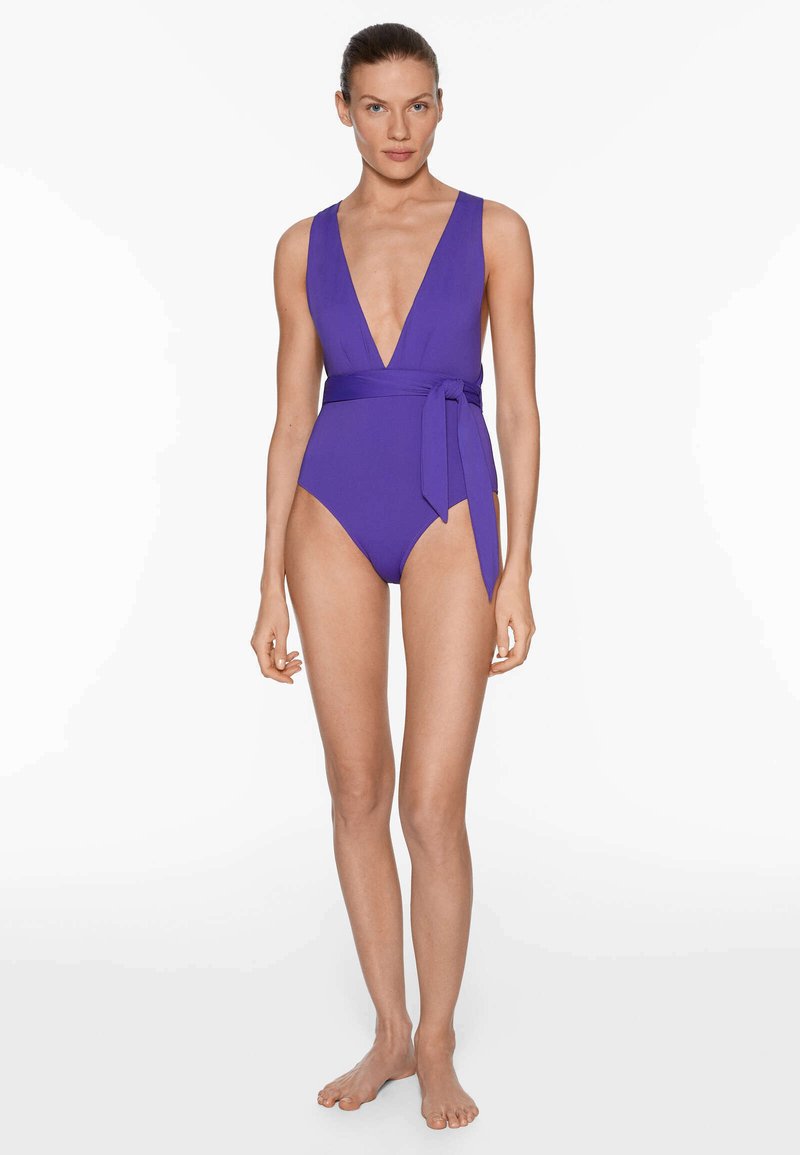 Maillot de bain une pièce violet avec un décolleté en V profond et un détail de lien noué à la taille. Texture lisse, design ajusté, jambes à taille haute.