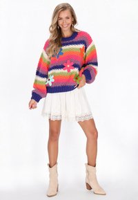 Mehrfarbiger gestrickter Pullover mit horizontalen Streifen, Blumen-Applikationen, langen Ärmeln, kombiniert mit einem weißen Rock mit Spitze und beigen Stiefeln.