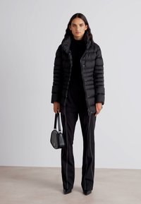 Zwarte pufferjack met verticale quiltontwerp, ritssluiting en een capuchon; gecombineerd met zwarte op maat gemaakte broek en een kleine handtas.