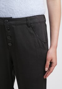 Pantaloni neri realizzati in un tessuto liscio, con primo piano di una chiusura a cinque bottoni, tasca laterale e vita piatta.