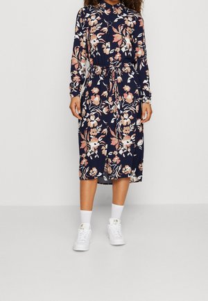 Woman wearing a navy blue floral midi dress with long sleeves, a waist tie, white socks, and white sneakers standing against a plain background.