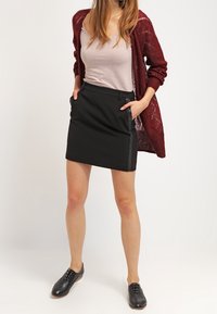 Falda negra ajustada con bolsillos laterales, combinada con una camiseta sin mangas rosa clara y un cárdigan de punto burdeos. Unos zapatos negros con cordones completan el conjunto.