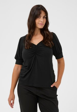 Une femme porte un haut noir à col V, froncé sur le devant et doté de manches bouffantes, associé à un pantalon noir sur un fond clair et uni.