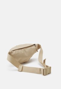 Béžová crossbody taška vyrobená z měkké látky s předním zipovým uzávěrem, nastavitelného popruhu a zaobleného designu. Vhodná pro každodenní použití.