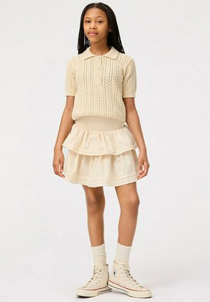 Beige stickad polo med korta ärmar, kombinerad med en lager-på-lager krämfärgad kjol. Vita högskaftade sneakers och knästrumpor fulländar outfiten.