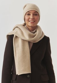 Beige strikket hue og matchende tørklæde, begge med en blød tekstur. Tørklædet er løst svunget omkring en neutralt tonet bluse og sort frakke.