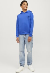 Modrá mikina s kapucí a černým potiskem na levé straně. Spárováno s lehce opotřebovanými světle modrými džíny a bílými teniskami.