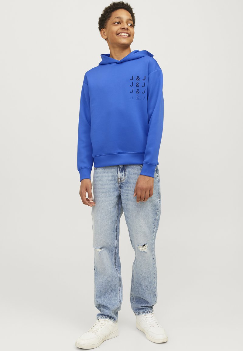 Modrá mikina s kapucí a černým potiskem na levé straně. Spárováno s lehce opotřebovanými světle modrými džíny a bílými teniskami.