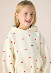 Crème hoodie met een buidelzak, versierd met verspreide kleine rode hartjespatronen, gemaakt van zachte stof met een ontspannen pasvorm en een capuchon.