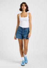 Blaue Denim-Shorts mit einem rohen Saum, kombiniert mit einem weißen gerippten Tanktop. Das Modell trägt hellblaue Sneakers und weiße Socken und steht vor einem grauen Hintergrund.