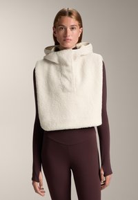 Witte, fluffy fleece crop top met een capuchon en een voorzak, gecombineerd met een strakke bruine lange mouw top en bijpassende legging.